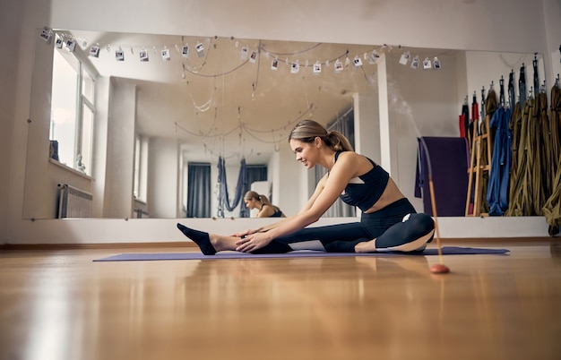 Attractive blond woman stretching her body while sitting on special rubber yoga mat in gym