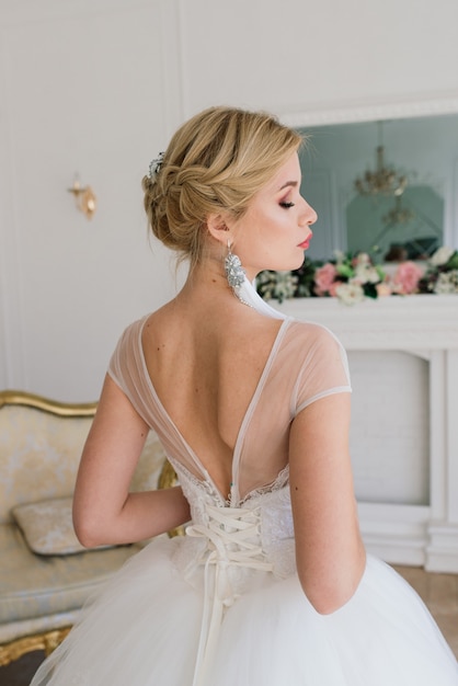 Attractive blonde bride is standing by the window in a white interior in vintage style.