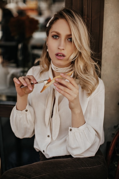 Attractive blonde curly woman spreads paste on bruschetta. Pretty lady in white blouse and velvet pants sits in street cafe