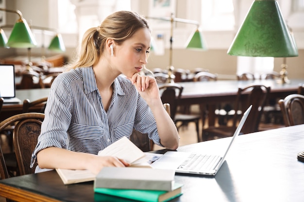 Attractive blonde girl student studying at the library with laptop computer