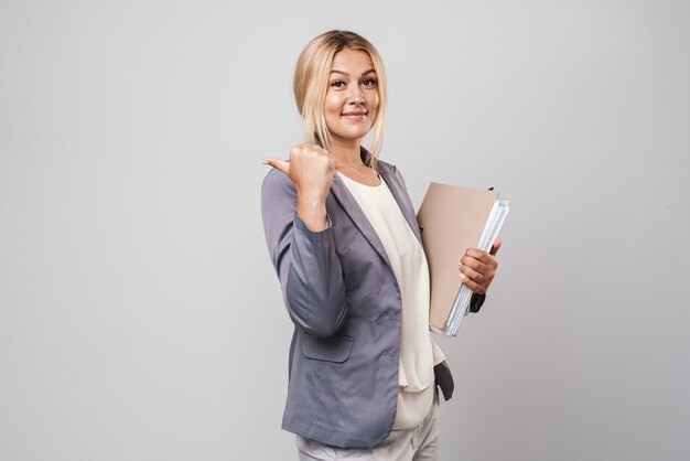 Attractive blonde haired businesswoman wearing jacket standing isolated over gray wall, carrying folders, pointing at copy space