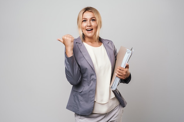 Attractive blonde haired businesswoman wearing jacket standing isolated over gray wall, carrying folders, pointing at copy space