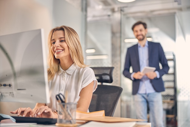 Attractive blonde lady working on desktop PC and smiling while male colleague standing behind her