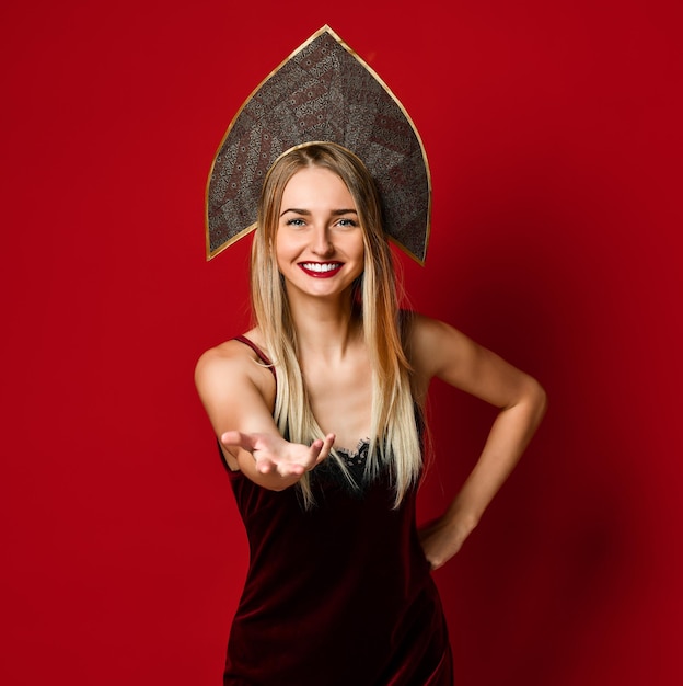 Attractive blonde woman in kokoshnik raised up palm arm hand at you isolated over red background