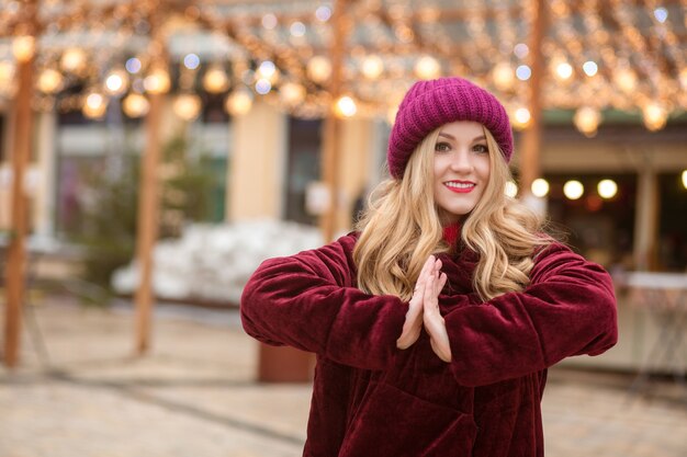 Attractive blonde woman wearing red knitted hat and winter coat, posing on the background of garland at the street in Kyiv