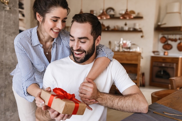 attractive brunette couple man and woman having breakfast in apartment while sitting at table with present box