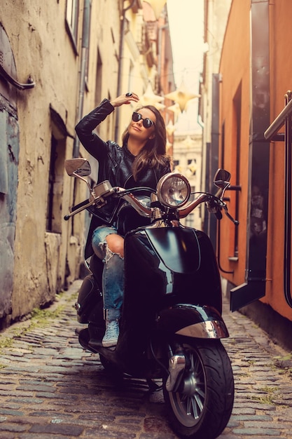 Attractive brunette female in leather jacket sitting on a motor scooter on a street in old town.