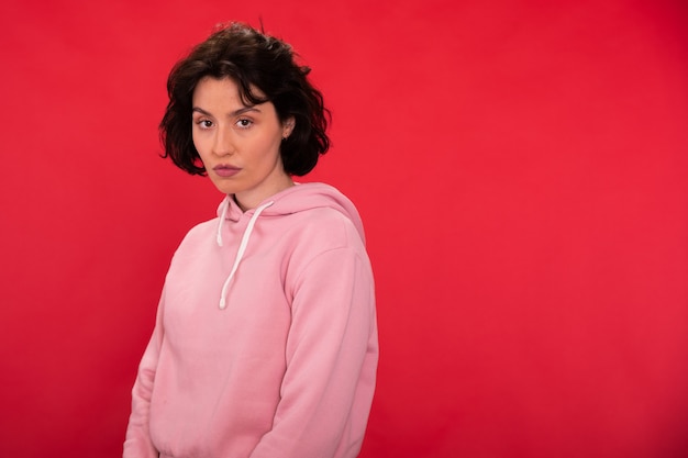 Attractive brunette girl in pink hoodie posing on red studio backdrop copy space