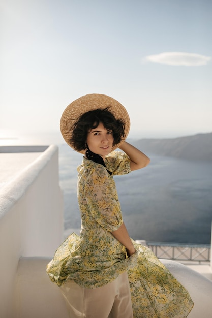 Attractive brunette woman in boater and floral dress looks into camera on balcony with sea view Shorthaired lady in hat smiles and poses on terrace on ocean background