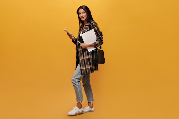 Attractive brunette woman in jeans and oversized jacket looks into camera moves holds phone and notebooks on yellow isolated background