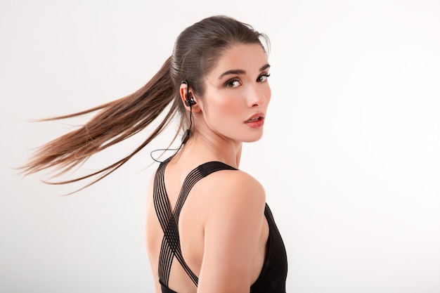 Attractive brunette woman in jogging black top listening to music on earphones posing isolated on white wall ponytail hairstyle waving long hair