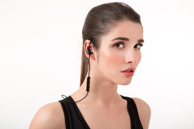 Attractive brunette woman in jogging black top listening to music on earphones posing isolated on white wall ponytail hairstyle