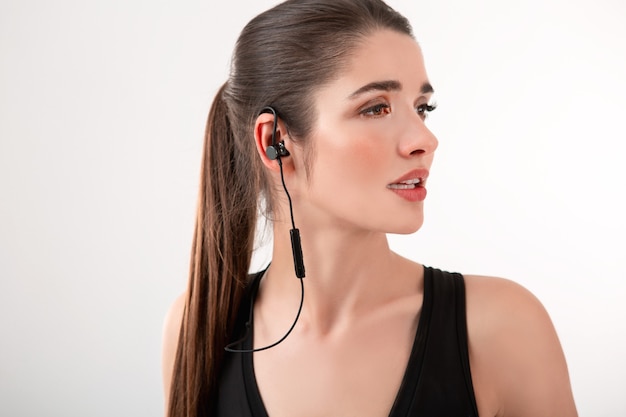 Attractive brunette woman in jogging black top listening to music on earphones posing isolated on white wall ponytail hairstyle