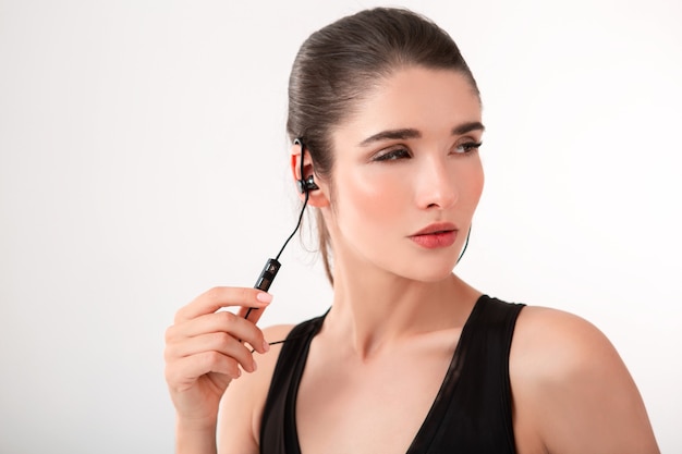 Attractive brunette woman in jogging black top listening to music on earphones posing isolated on white wall