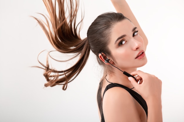 Attractive brunette woman in jogging black top listening to music on earphones posing isolated on white wall