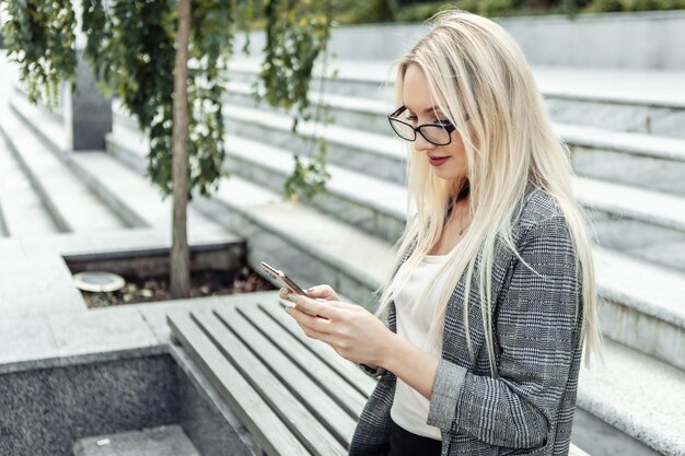 Attractive business woman holding smartphone in her hands while sitting on bench outdoors