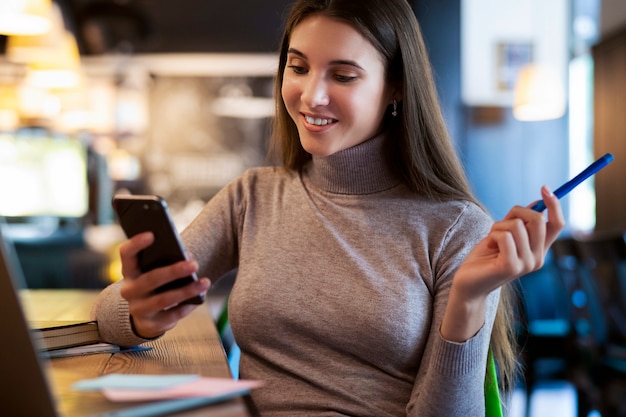 Attractive business woman sits at table in front of laptop, holds smartphone, pencil in her hand.