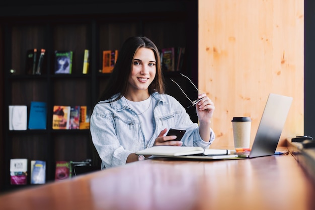 Attractive business woman sits at table in front of laptop, writes message, uses smartphone, talks on phone.