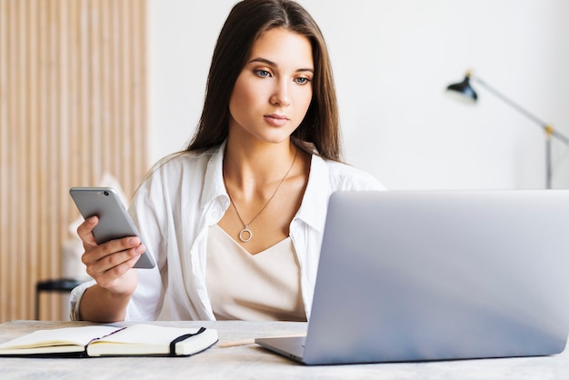 Attractive business woman sits at table in laptop, smartphone lies on table, makes notes with pencil in notebook.