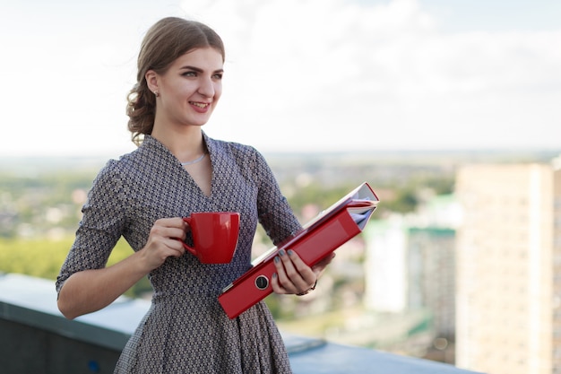 Attractive businesslady in patterned dress stand on the roof and hold paper folder and red cup