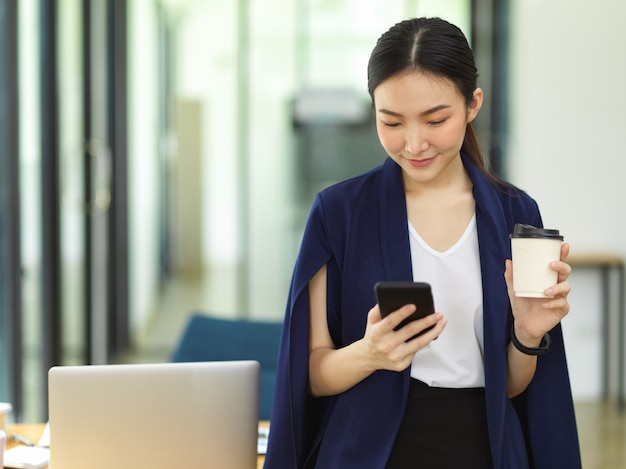 Attractive businesswoman drinking a coffee and using a smart mobile phone while standing in office, Young business entrepreneur message with business supplier in office