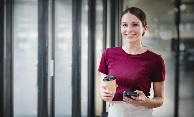 Attractive businesswoman holding a coffee and mobile phone while standing at a window in an office smiling and looking at camera