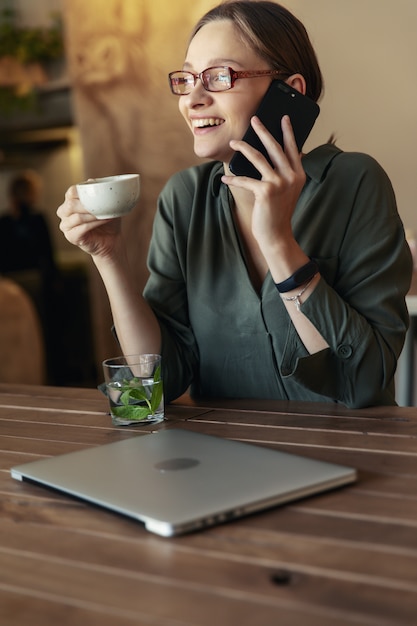 Attractive  businesswoman in jacket and stylish glasses sitting in cafe, talking on the phone and looking at laptop.