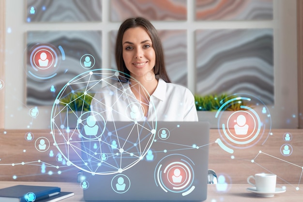 Attractive businesswoman in white shirt at workplace working with laptop to hire new employees for international business consulting HR social media hologram icons over office background