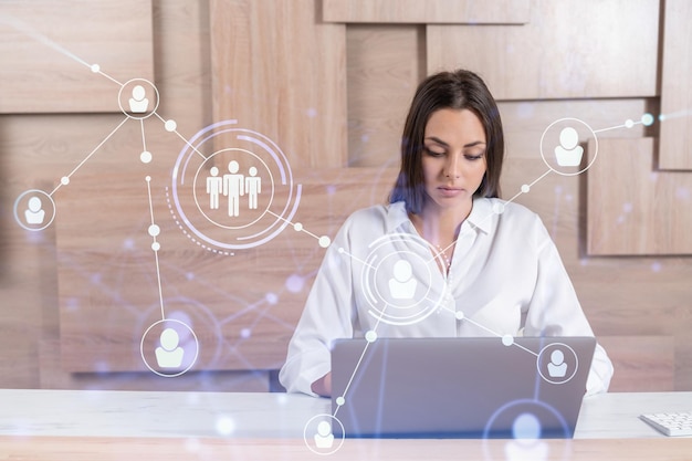 Attractive businesswoman in white shirt at workplace working with laptop to hire new employees for international business consulting HR social media hologram icons over office background