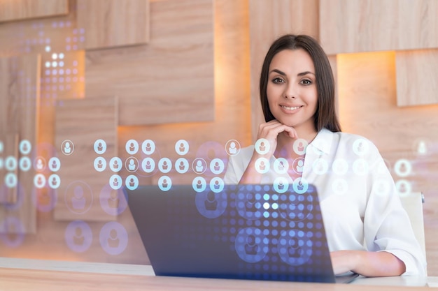 Attractive businesswoman in white shirt at workplace working with laptop to hire new employees for international business consulting HR social media hologram icons over office background