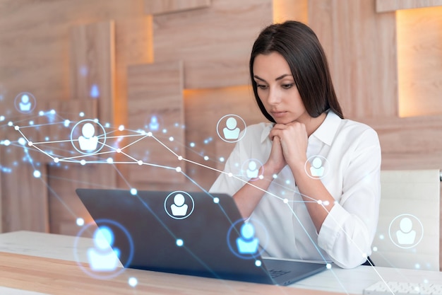 Attractive businesswoman in white shirt at workplace working with laptop to hire new employees for international business consulting HR social media hologram icons over office background