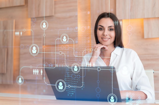 Attractive businesswoman in white shirt at workplace working with laptop to hire new employees for international business consulting HR social media hologram icons over office background