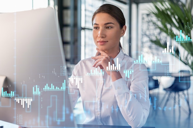 Attractive businesswoman in white shirt at workplace working with laptop to optimize trading strategy at corporate finance fund Forex chart hologram over office background