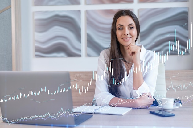 Attractive businesswoman in white shirt at workplace working with laptop to optimize trading strategy at corporate finance fund Forex chart hologram over office background