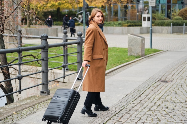 Attractive casual girl in beige coat walking around city street with suitcase