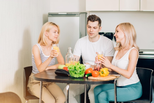 Attractive caucasian blonde girl in casual clothes sitting with friends in kitchen, looking at camera with smile. Selective focus. People, healthy food concept.