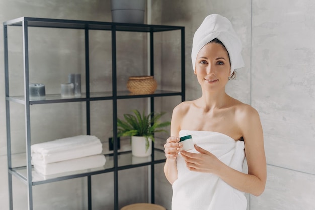 Attractive caucasian girl wrapped in towel after bathing and hair washing is holding a cream jar