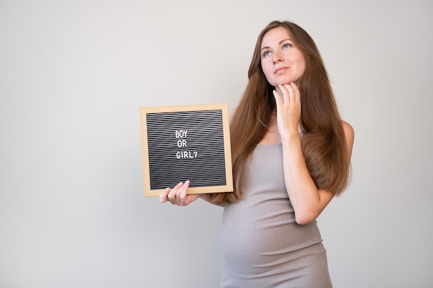 Attractive caucasian pregnant thoughtful dreaming woman holding board with boy or girl question on light gray background Gender reveal conceptCopy space for text