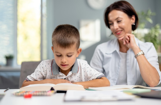 Attractive caucasian woman sitting at desk with his son and reading book Schoolboy doing homework with caring mother Concept of parenting and support