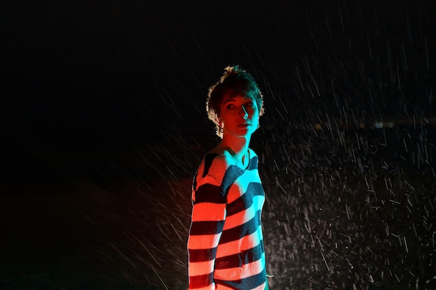 Attractive Caucasian woman stands on evening rainy street in light of car headlights and looks back