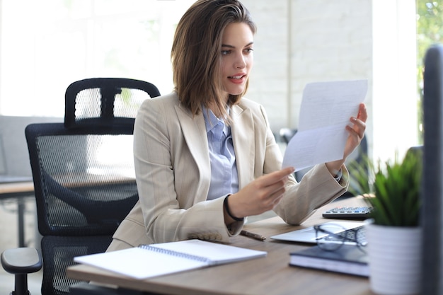 Attractive cheerful business woman checking paper documents in office, working on laptop.