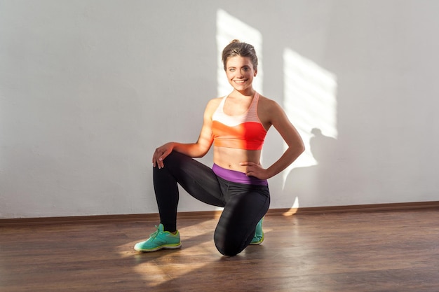 Attractive cheerful sportive woman with bun hairstyle and in tight sportswear standing on one knee and smiling at camera resting after workouts indoor studio shot illuminated by sunlight from window