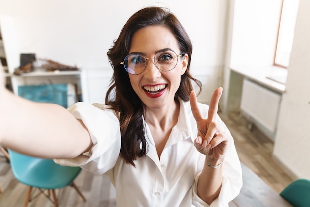 Attractive cheerful young brunette businesswoman standing in the cafe indoors, taking a selfie