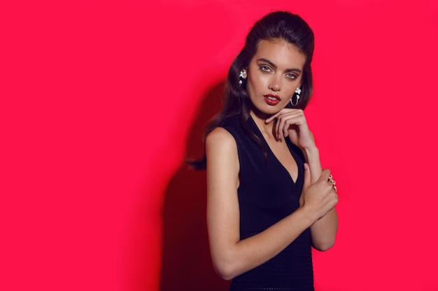 Attractive, cheerful young woman with makeup and hairstyle, red lipstick in elegant slim black dress, wears jewellery, over red background.