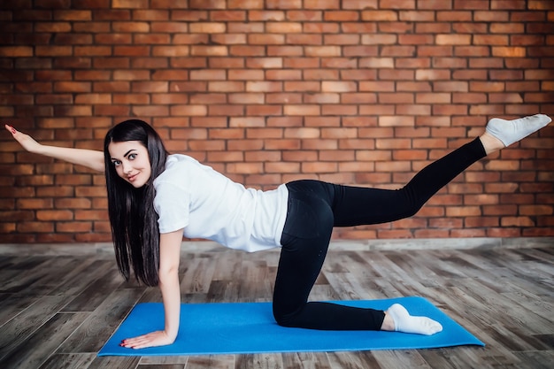 Attractive cheerful young woman working out indoors. Beautiful brunette  model doing exercises on blue mat in room with  walls.