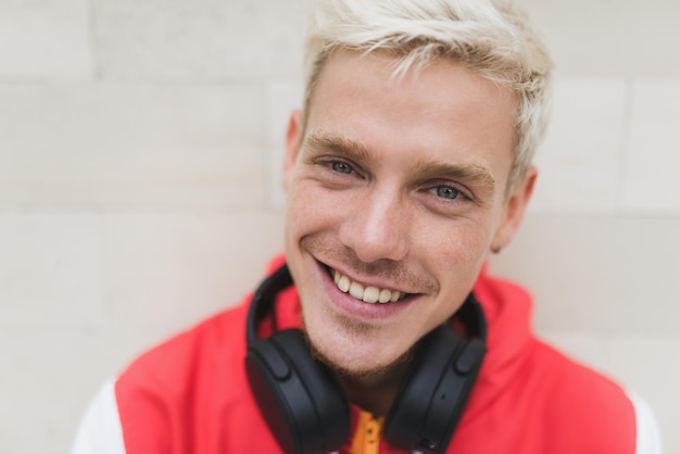 Attractive close up portrait of handsome young blond Caucasian man smiling and looking at the camera with headphones on light grey outdoor background Lifestyle people technology and music concept