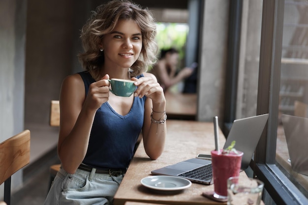 Attractive confident successful female entrepreneur sit cafe near window work laptop drink coffee hold cup smiling happily relaxed camera studying coworking space prepare project online