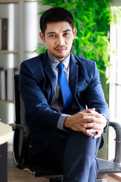 Attractive confident young Asian businessman sits on chair with leg crossed in office. Business and success concept.