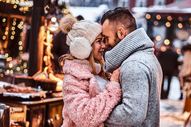 An attractive couple in love, a stylish couple wearing warm clothes cuddling together and looking each other at the winter fair at a Christmas time.