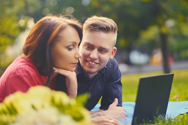 Attractive couple lying on a blanket on a lawn and is using a laptop on a picnic.